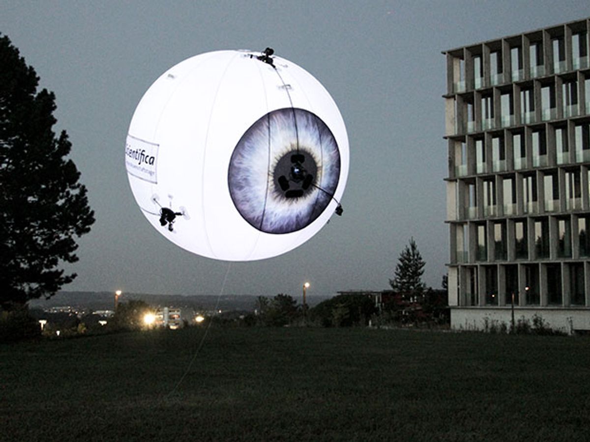 A glowing floating balloon patterned to look like an eyeball hovers in the evening sky outdoors.
