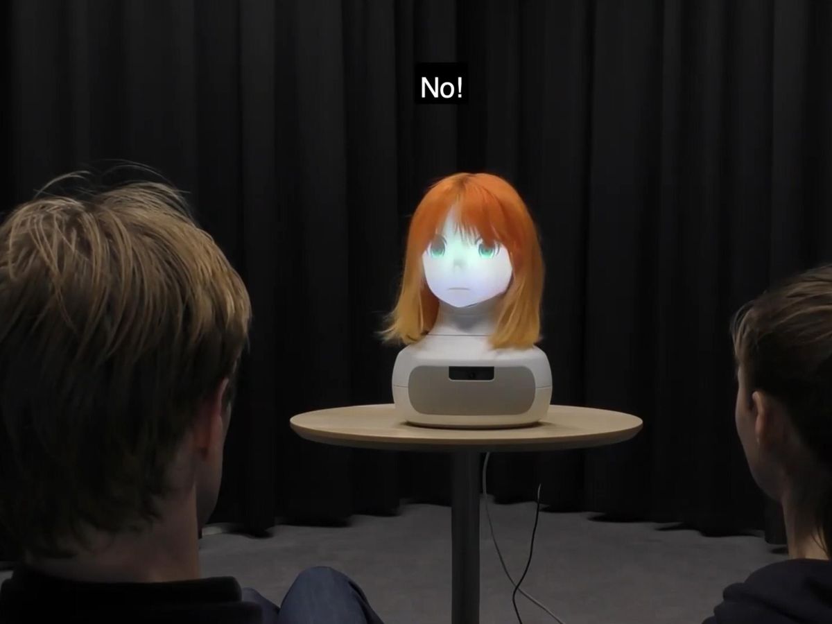 A glowing robot face with big green eyes and a red wig is attached to a beige base sitting on a table. In the foreground are the backs of a male (left) and female (right). Above the robot is "No!" and she appears to be looking at the male.