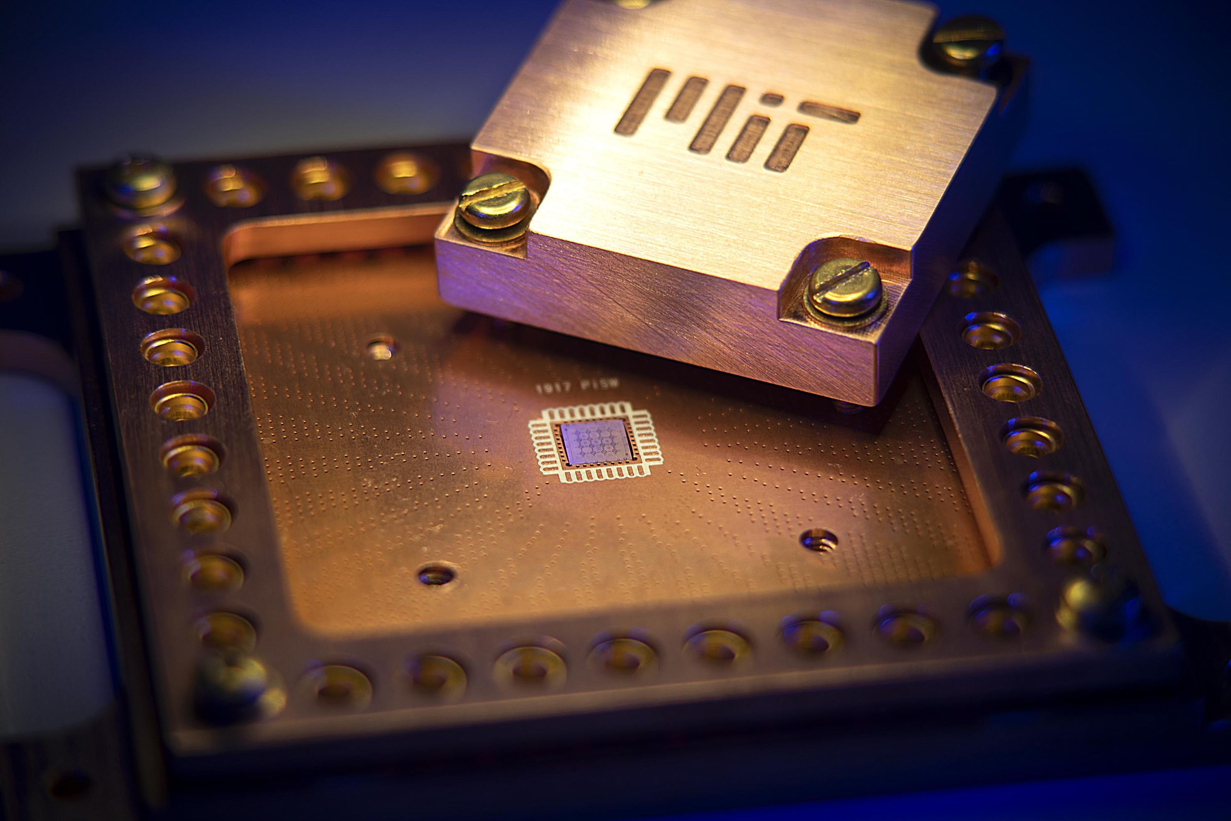 A golden square package holds a small processor. Sitting on top is a metal square with  MIT etched into it.