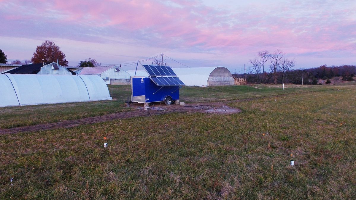 一片草地，地面上散落着白色的盒子，一辆装有太阳能电池板的蓝色拖车，以及多个白色的温室帐篷。 