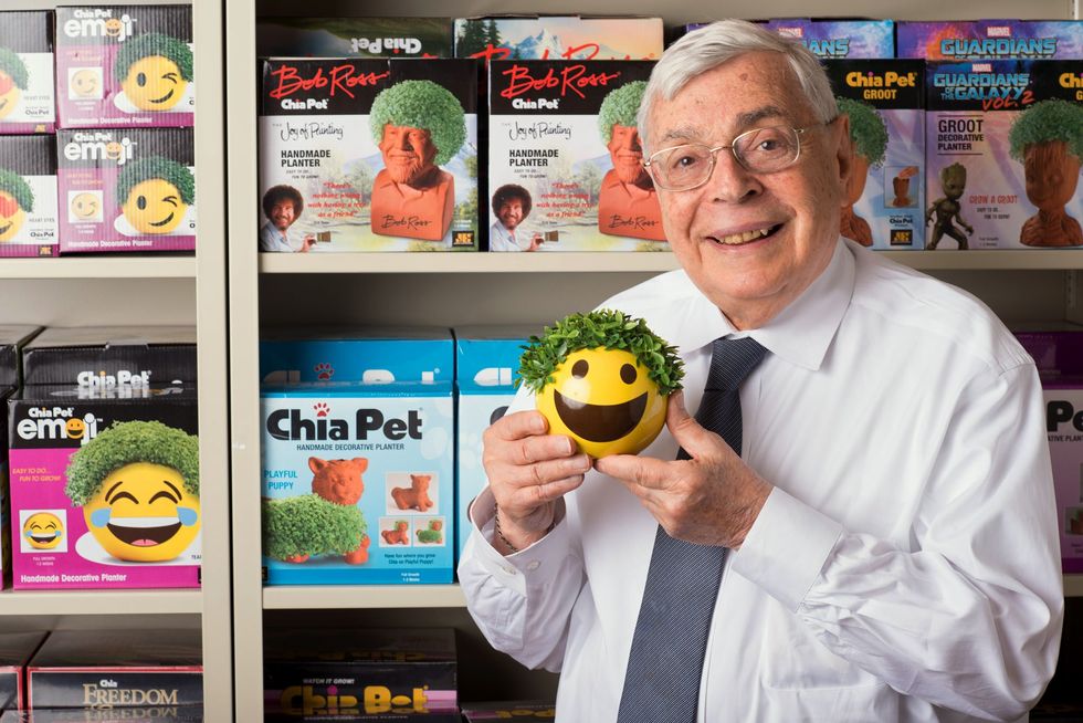 A gray haired bespectacled man in a white shirt and gray tie smiles at the camera. He is standing in front of shelves containing a variety of boxes for different Chia Pets, and is holding a smiley face emoji Chia Pet with a head of green leaves.