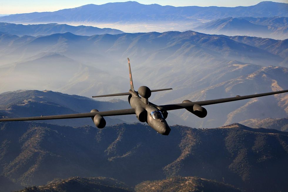 A gray military plane with wide wings flies over a mountainous landscape.