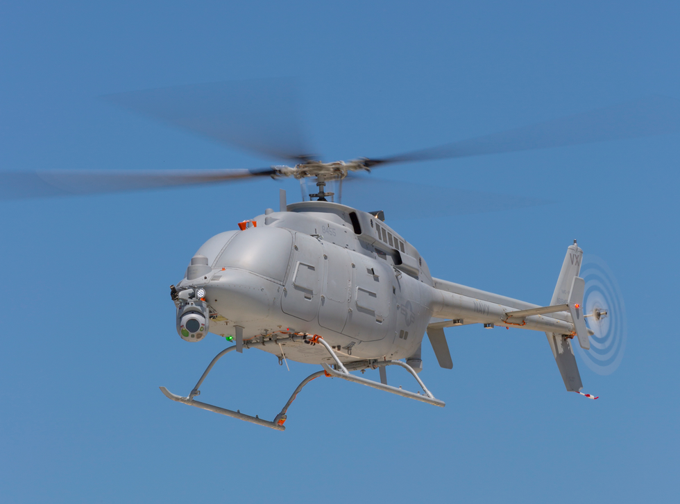 A gray unmanned helicopter, notably lacking a cockpit or any kind of window, is shown hovering against a clear, blue sky. It carries a downward-poinging sensor under its nose.
