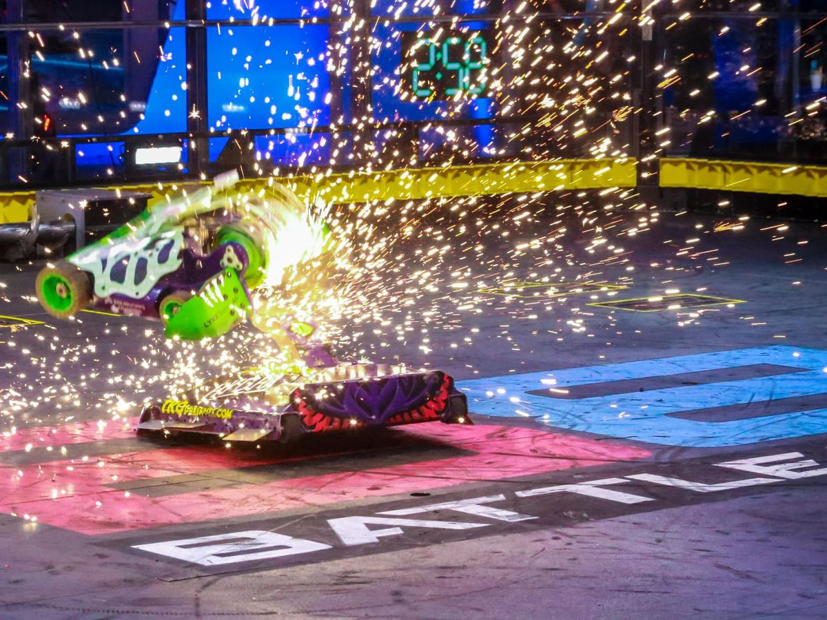 A green and purple Battlebot hovers in the air amid a large number of sparks. Below it is a purple and red Battlebot.