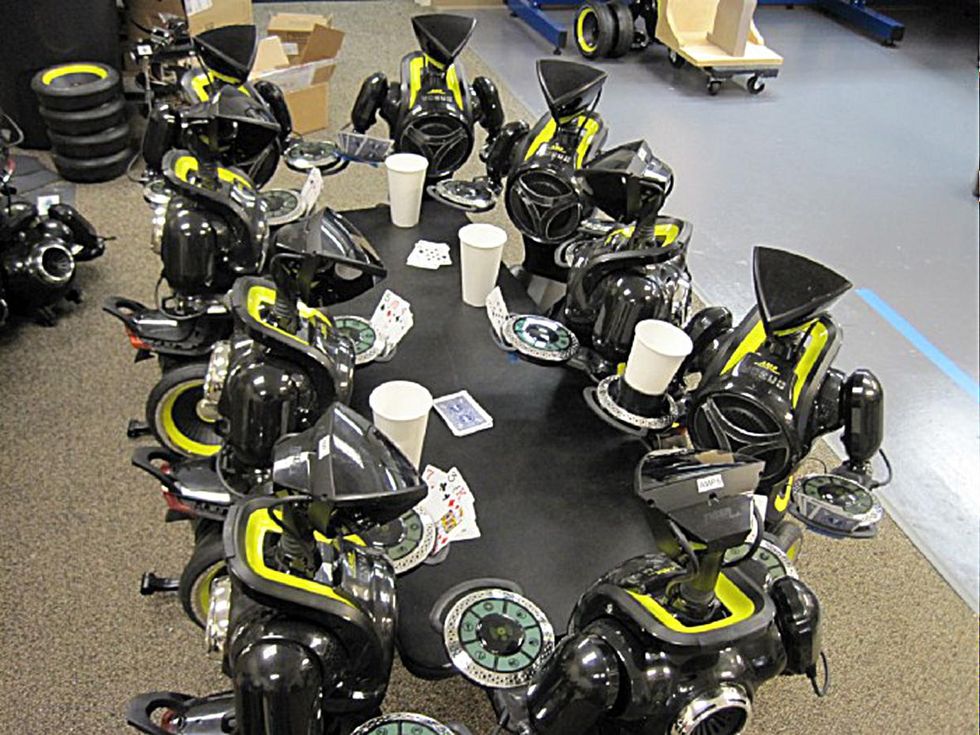 A group of AMP robots surround a black table holding drinks and playing cards