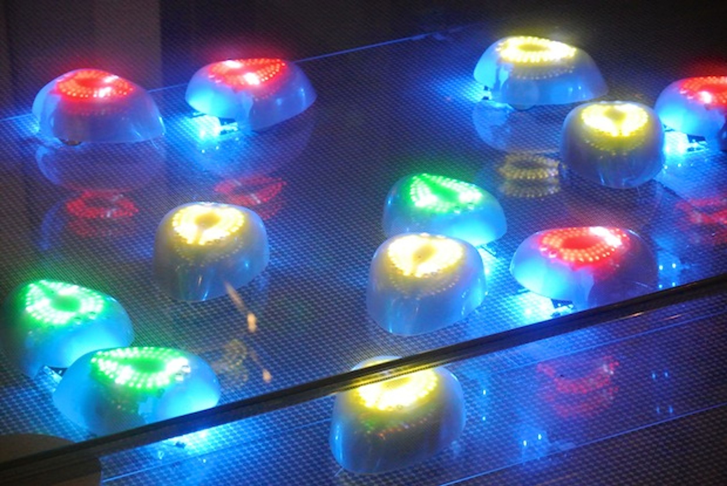 A group of ovoid robots underneath a transparent floor, illuminated by yellow, green, or red LEDs on their backs.