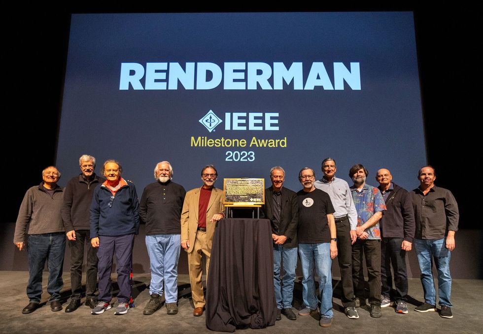 A group of people standing next to a plaque on a table with a screen projected behind them that reads RenderMan IEEE Milestone Award 2023