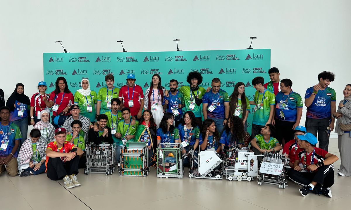 A group of students and adults from different countries pose with robots in front of signage for the First Global robotics competition.