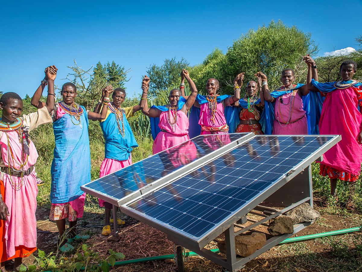 A group of women in a remote village in Leparua, Kenya hold hands in the air, surrounding a solar panel.
