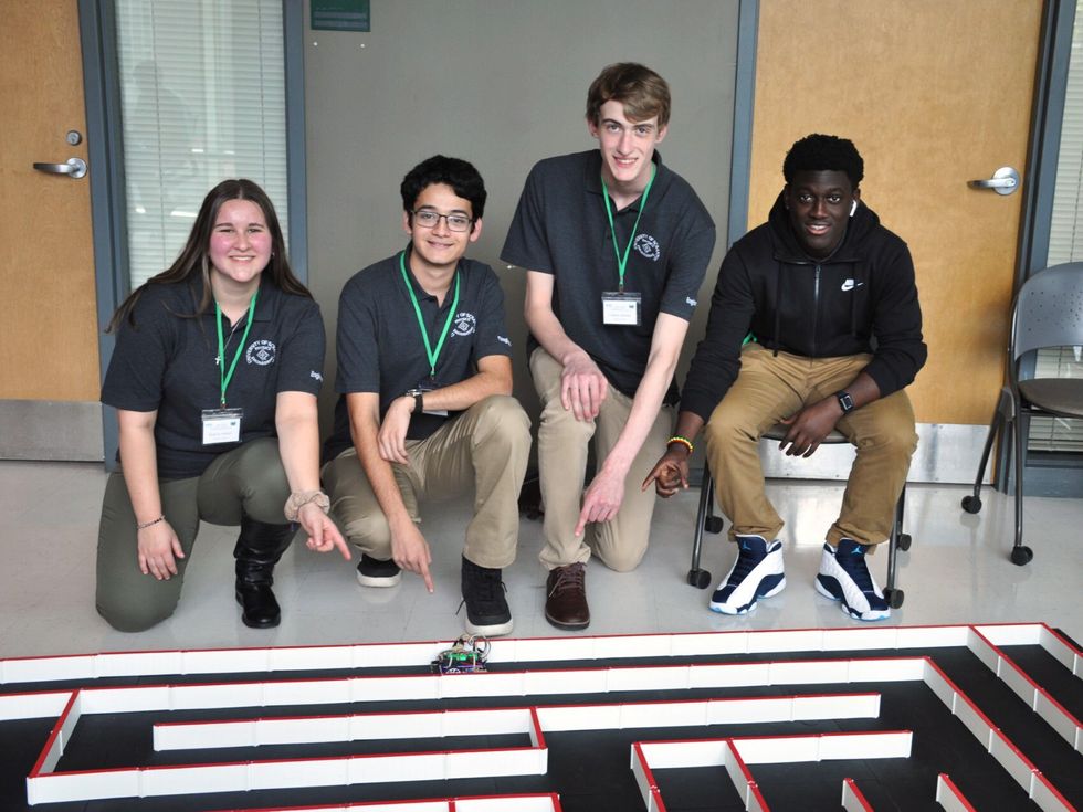 Group of young people kneeling in front of a maze-like structure on the ground and pointing at a small robot