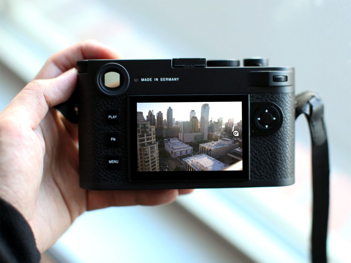 A hand holding a digital camera with a photograph of a city landscape on the screen. A small "cr" icon is at the right side of the image. 