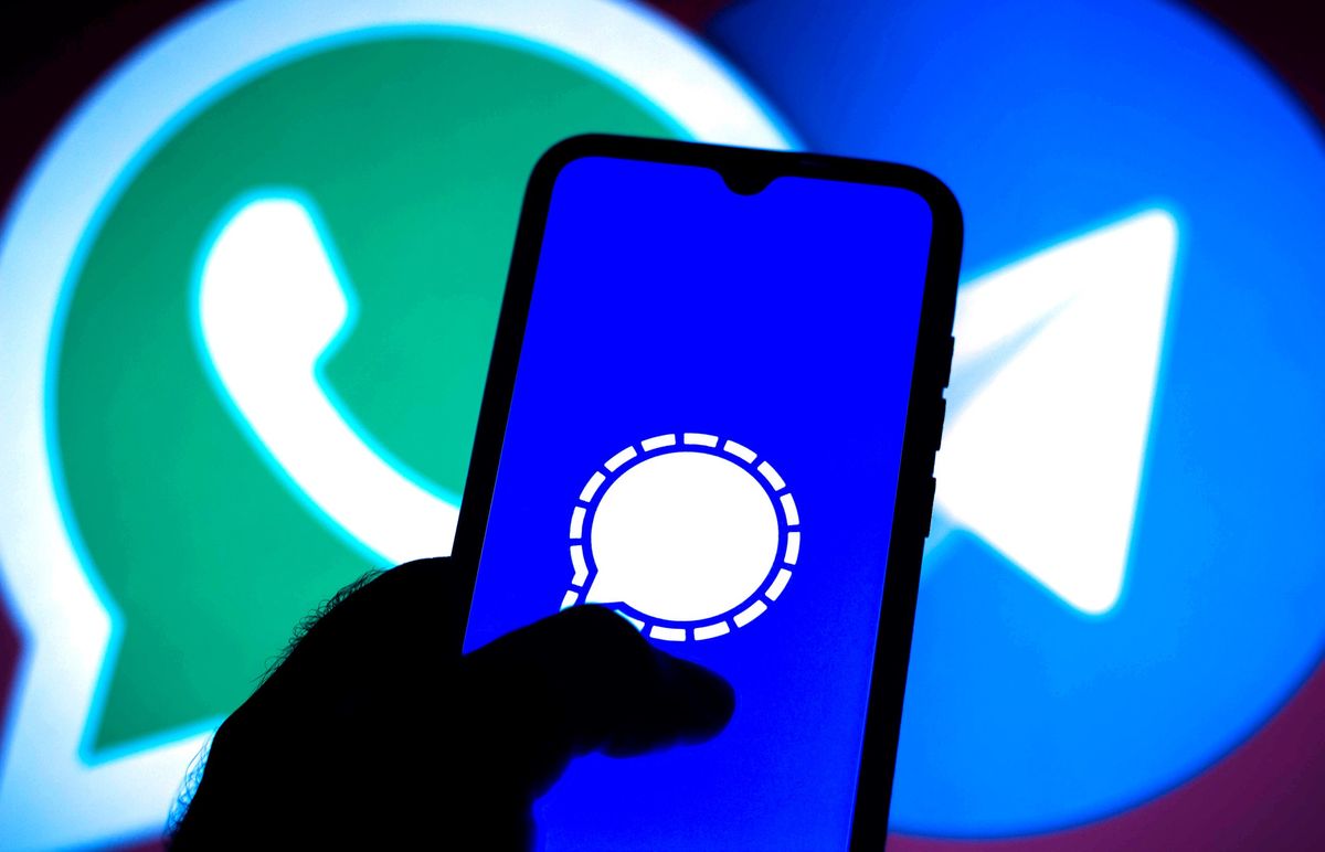 A hand holds a phone with the Signal logo in front of a display with the logos of Whatsapp and Telegram