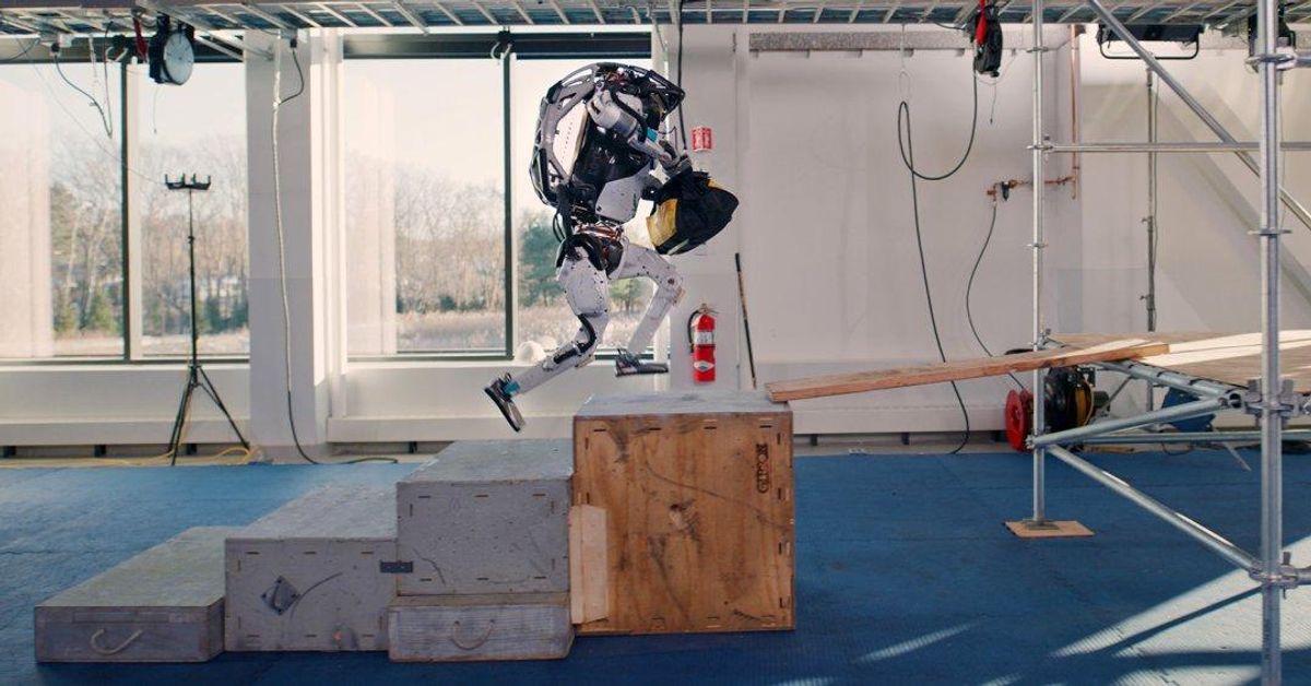 A humanoid robot holding a bag runs up a flight of stairs make of wooden boxes and approaches a wooden plank across a gap