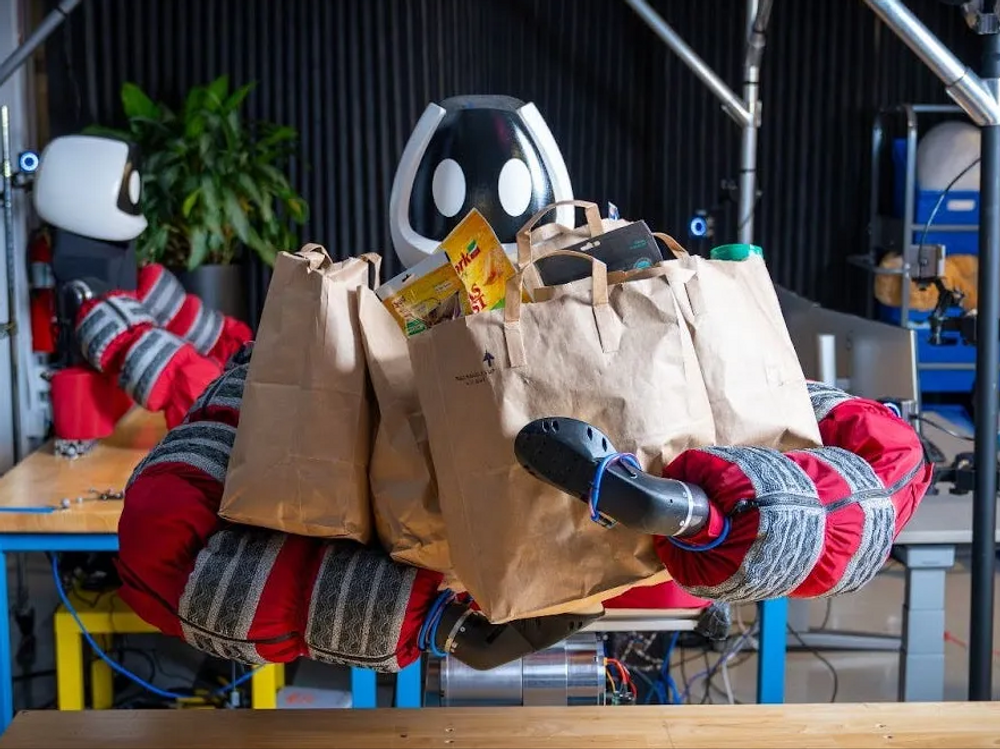 A humanoid robot with soft puffy sleeves grips several full grocery bags to its chest.