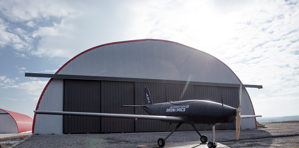 A large, black, propeller-driven drone stands in front of a warehouse 