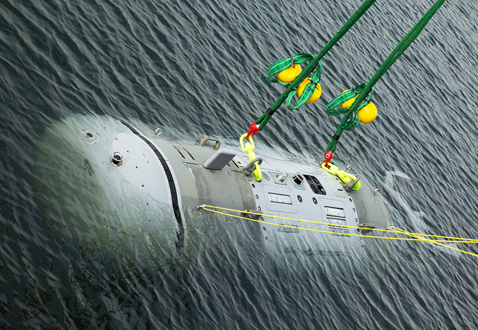 A large, cylindrical vehicle is shown just as it has been lowered below the surface of the water, suspended by two green cables.