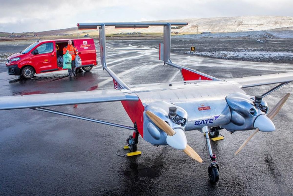 A large drone with twin propellers stands idle on a remote airport runway as a postal worker walks towards it with two large mail bags