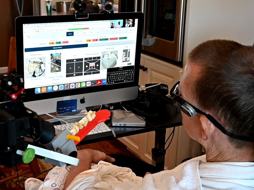 A large monitor shows an interface consisting of multiple views from cameras, simple maps of a house, and a keyboard. A man is seated in front of the screen, with the arm of a robot just visible, holding a kebab on a red flat tool.