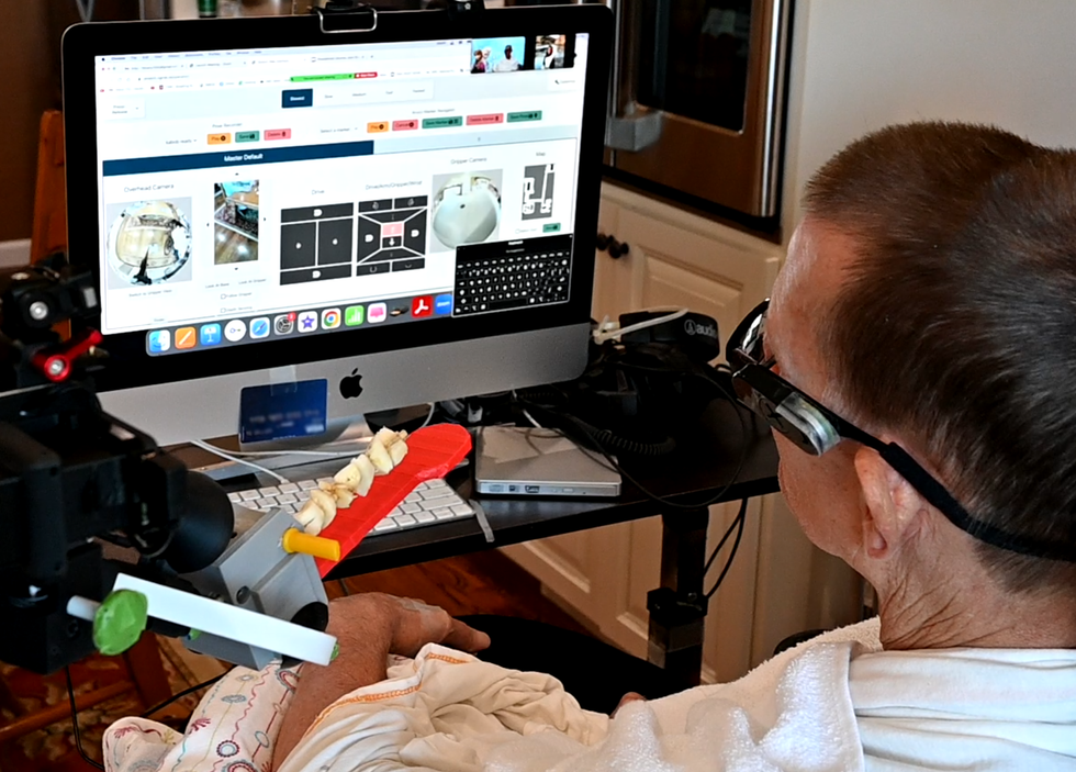 A large monitor shows an interface consisting of multiple views from cameras, simple maps of a house, and a keyboard. A man is seated in front of the screen, with the arm of a robot just visible, holding a kebab on a red flat tool.