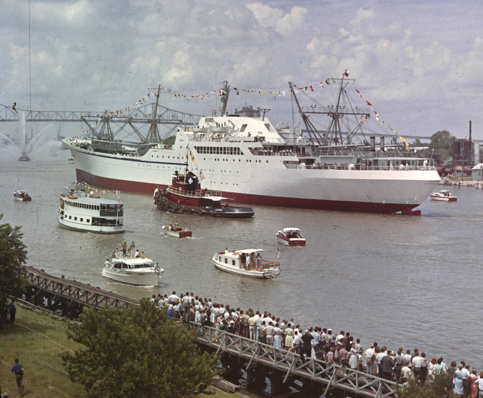 A large ship is shown near a bridge with a tugboat and several other smaller ships nearby.