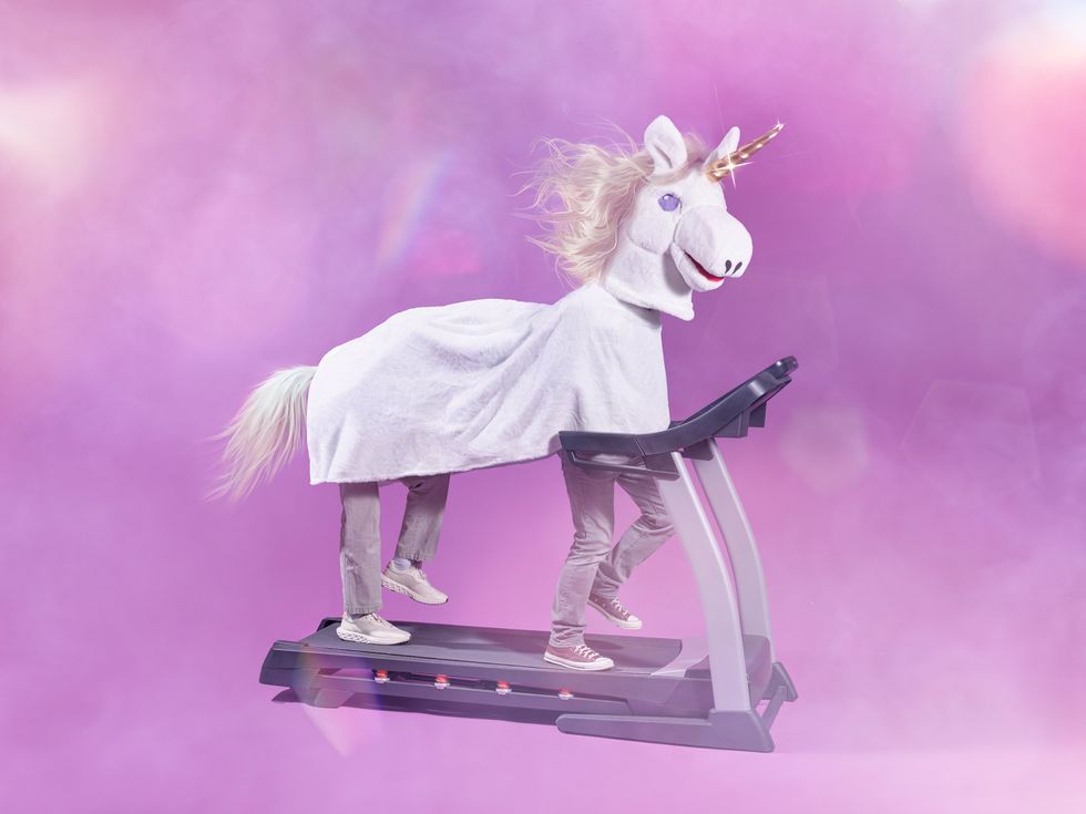 A large unicorn costume looks at the camera. Two people's legs are visible from inside the costume, and are moving on a treadmill.