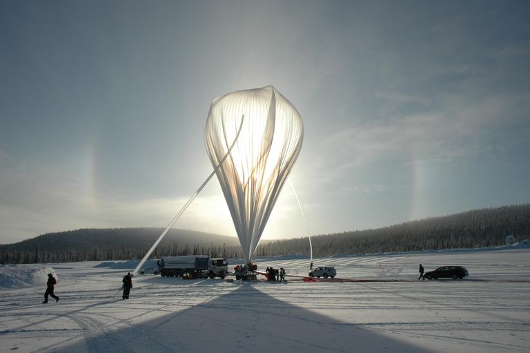 A large white scientific balloon is held by two people, with several vehicles near the base.