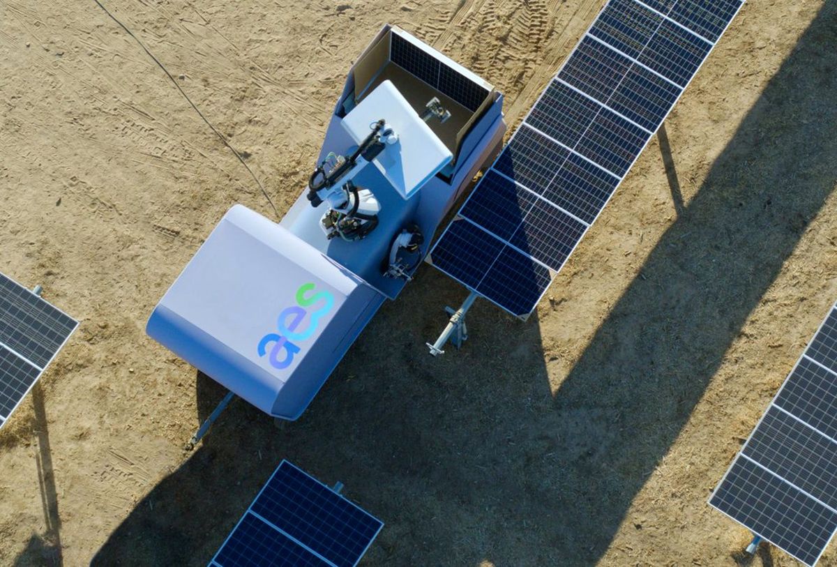 A large white wheeled robotic vehicle with a robotic arm next to solar panels in a field.