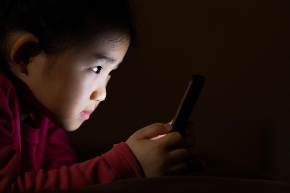 A little girls face lit up by the smart phone she is looking at in a dark room.