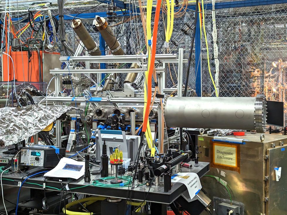 A long silver cylinder covered in equipment and wires rises from a work bench in a crowded lab.