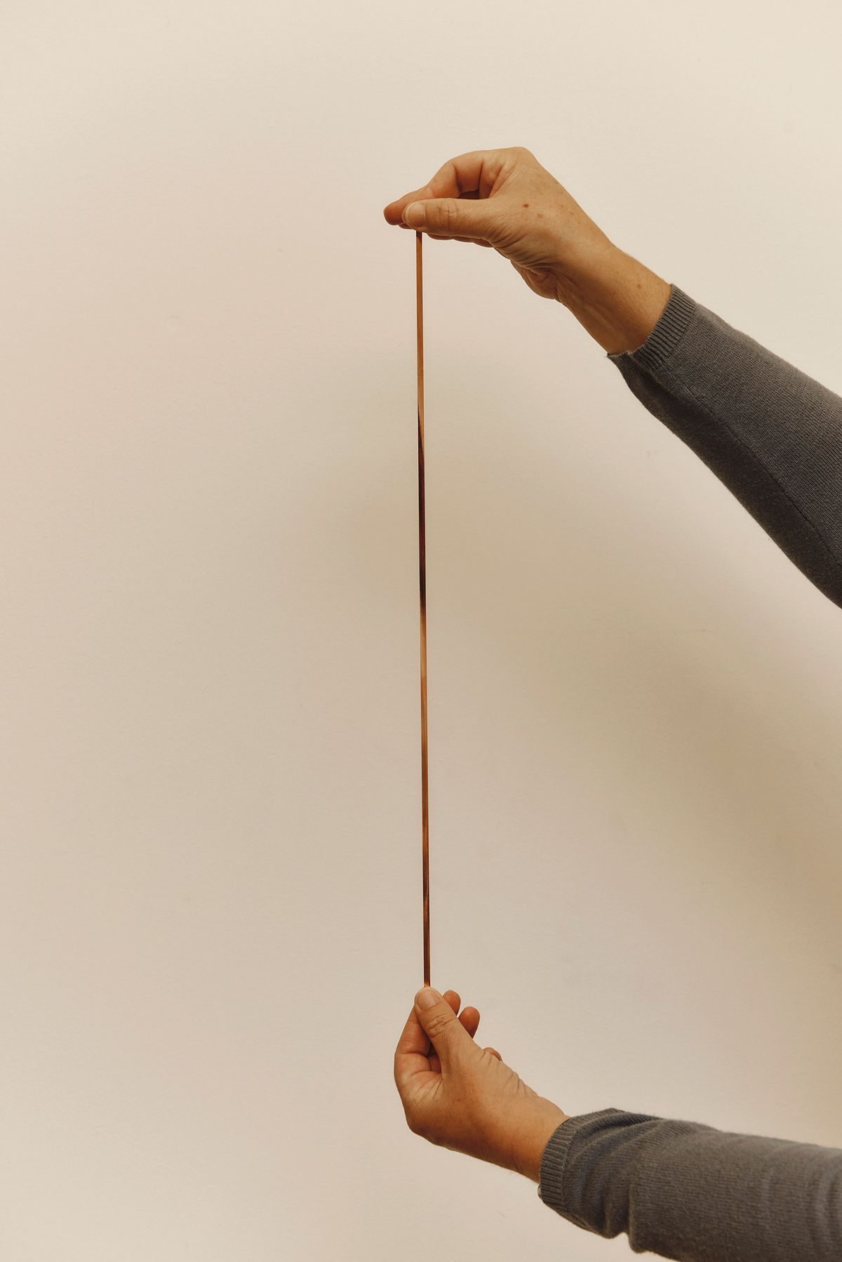 A long thin strip of coppery material is held vertically between a pair of hands in front of a beige background.