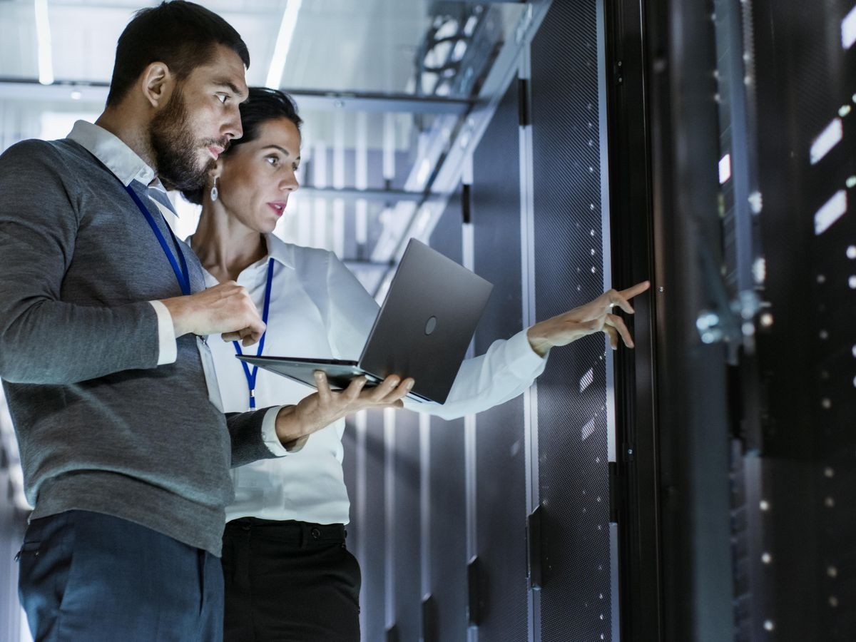 A man and woman in a data center. He holds a laptop while they confer.