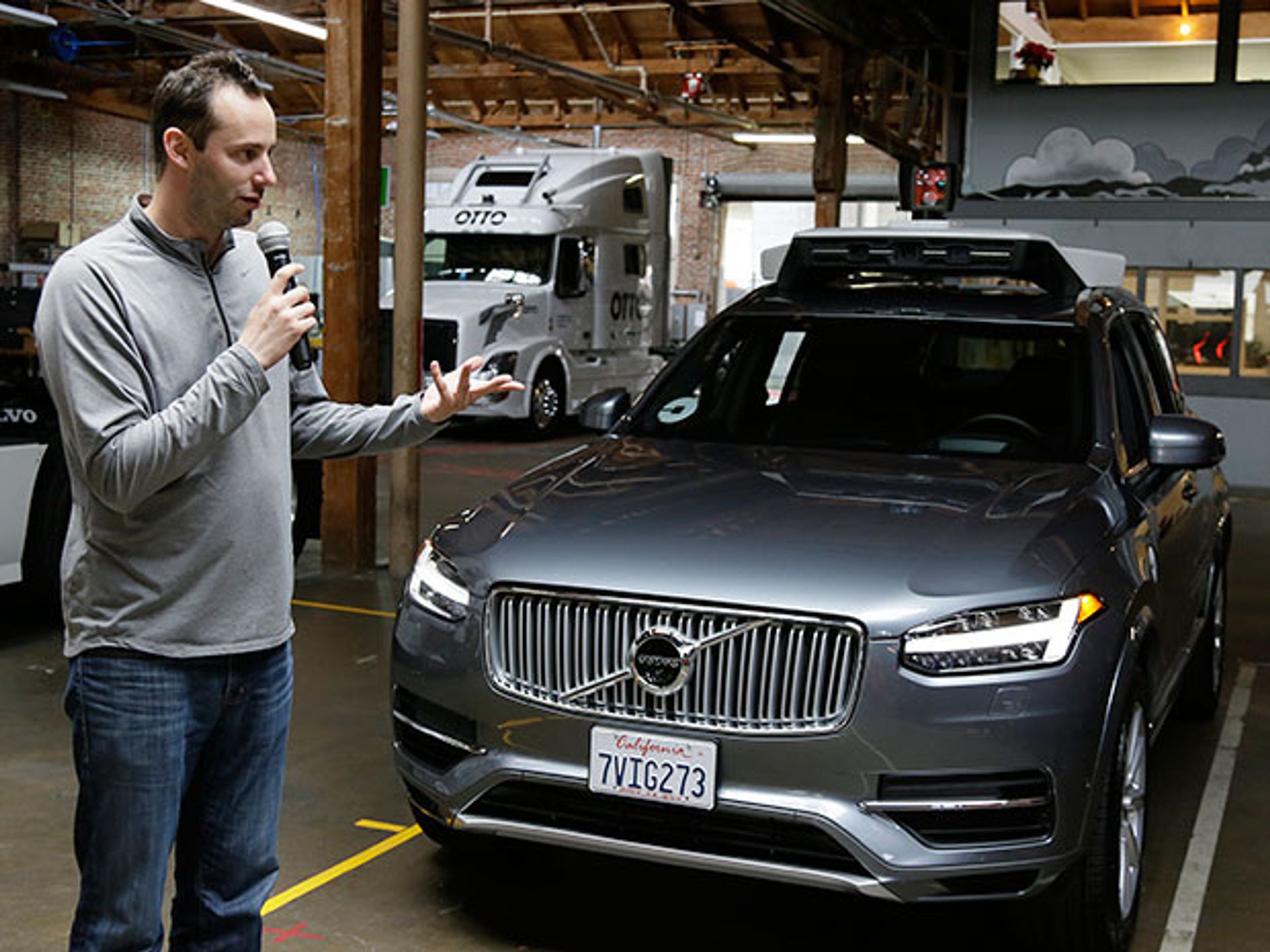 A man, Anthony Levandowski, in a gray shirt holding a microphone speaking in front of a silver sedan.