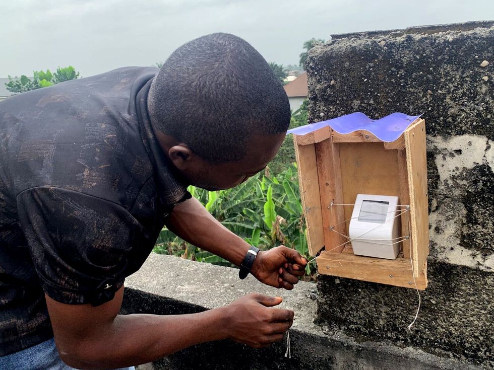 A man bends over as he secures a white sensor device to a wooden box with twine. The box is affixed to a stone fence outdoors.