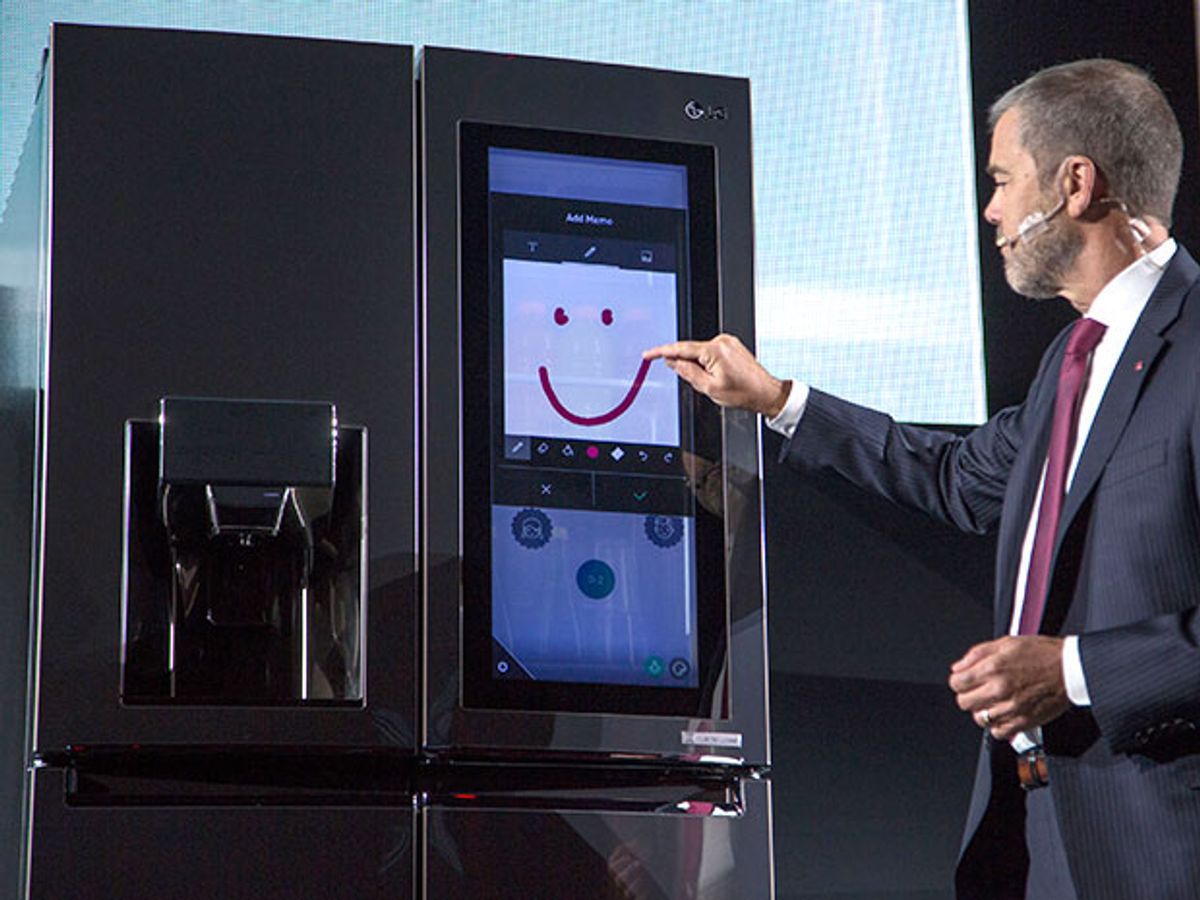 A man draws a crude smiley face on a touchscreen mounted on a gleaming metal fridge with french doors