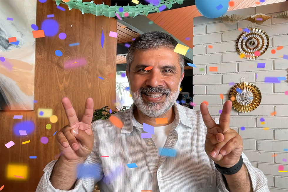 A man facing a camera has both hands raised and making a peace sign with two fingers on each hand. This triggers a burst of confetti to appear while using the FaceTime app in Apple iOS 17.