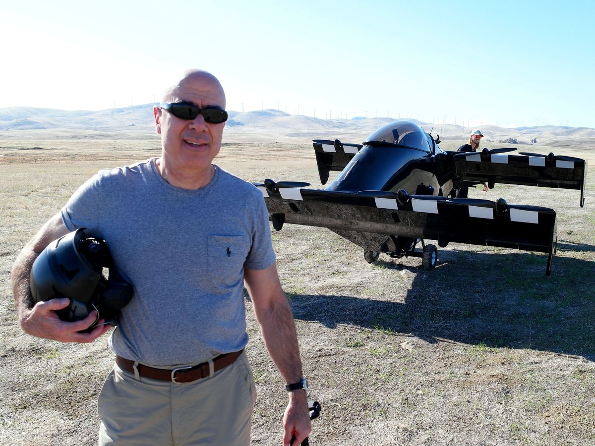 A man holding a helmet stands in front of a black eVTOL in an empty landscape.