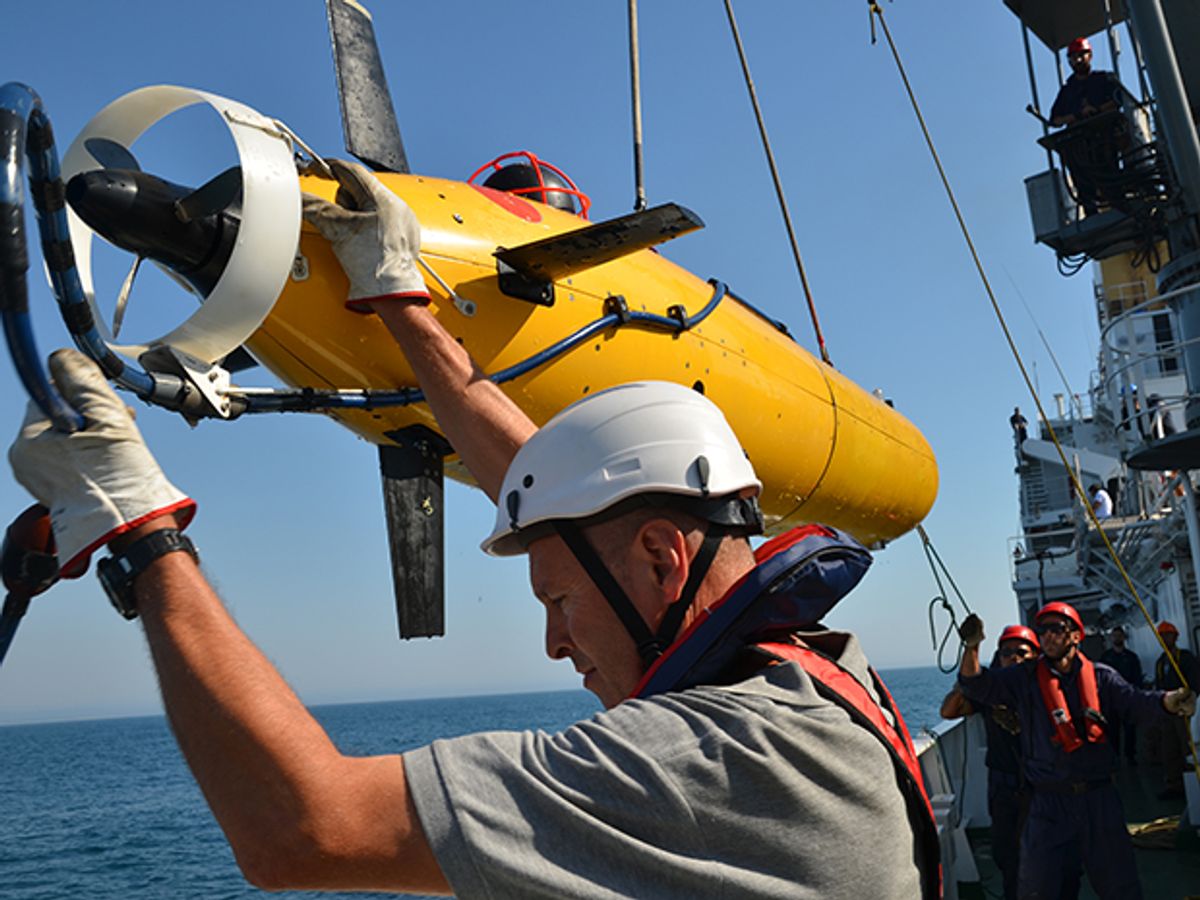 A man in a helmet lowers a yellow torpedo-shaped submersible over the side of a boat