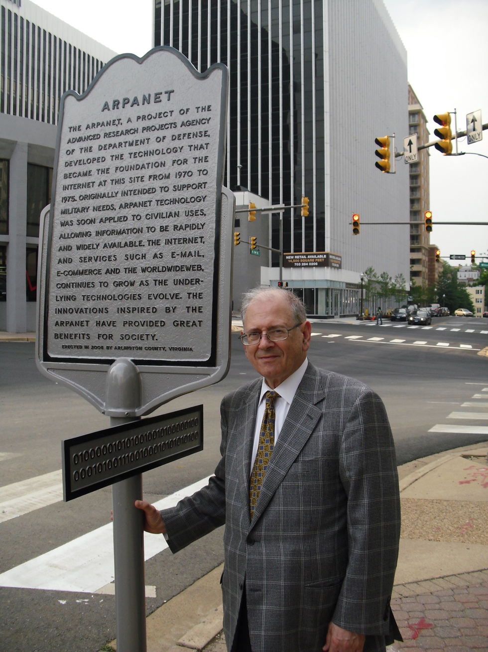A man in a suit stands in front of a sign with a paragraph of text.  The title, \u201cARPANET\u201d is legible.
