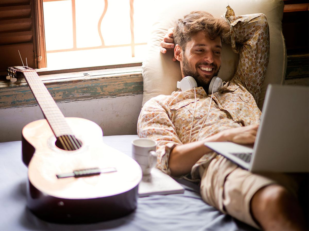 A man looks at his computer. A guitar sits next to him.