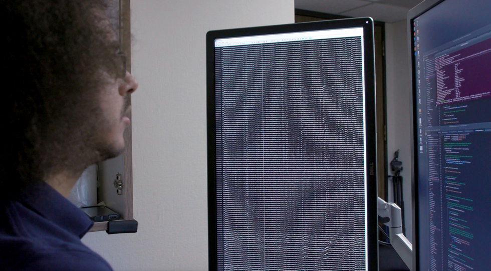 A man looks at two large display screens; one is covered in squiggly lines, the other shows text.\u00a0