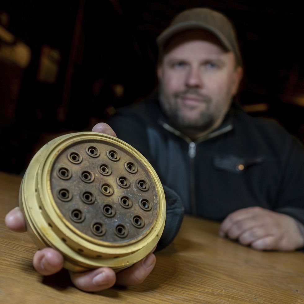 A man seated at a table holds a circular brass object toward the camera. The brass object has 19 large holes and has black char marks across its front. 