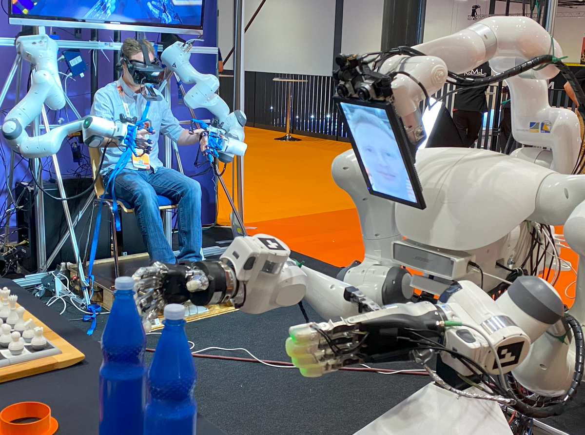 A man sits in a chair wearing VR googles and attached to two robotic arms. Near him, a robot with two arms show's the man's face on a screen while its two arms reach out for two blue bottles.