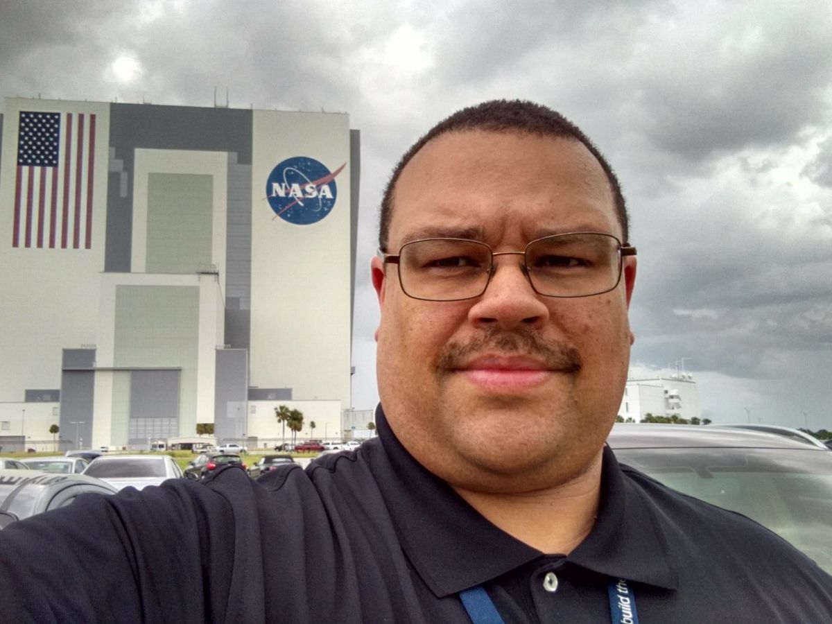 A man standing in a parking lot with cars and a building with the NASA logo and an american flag on it.  