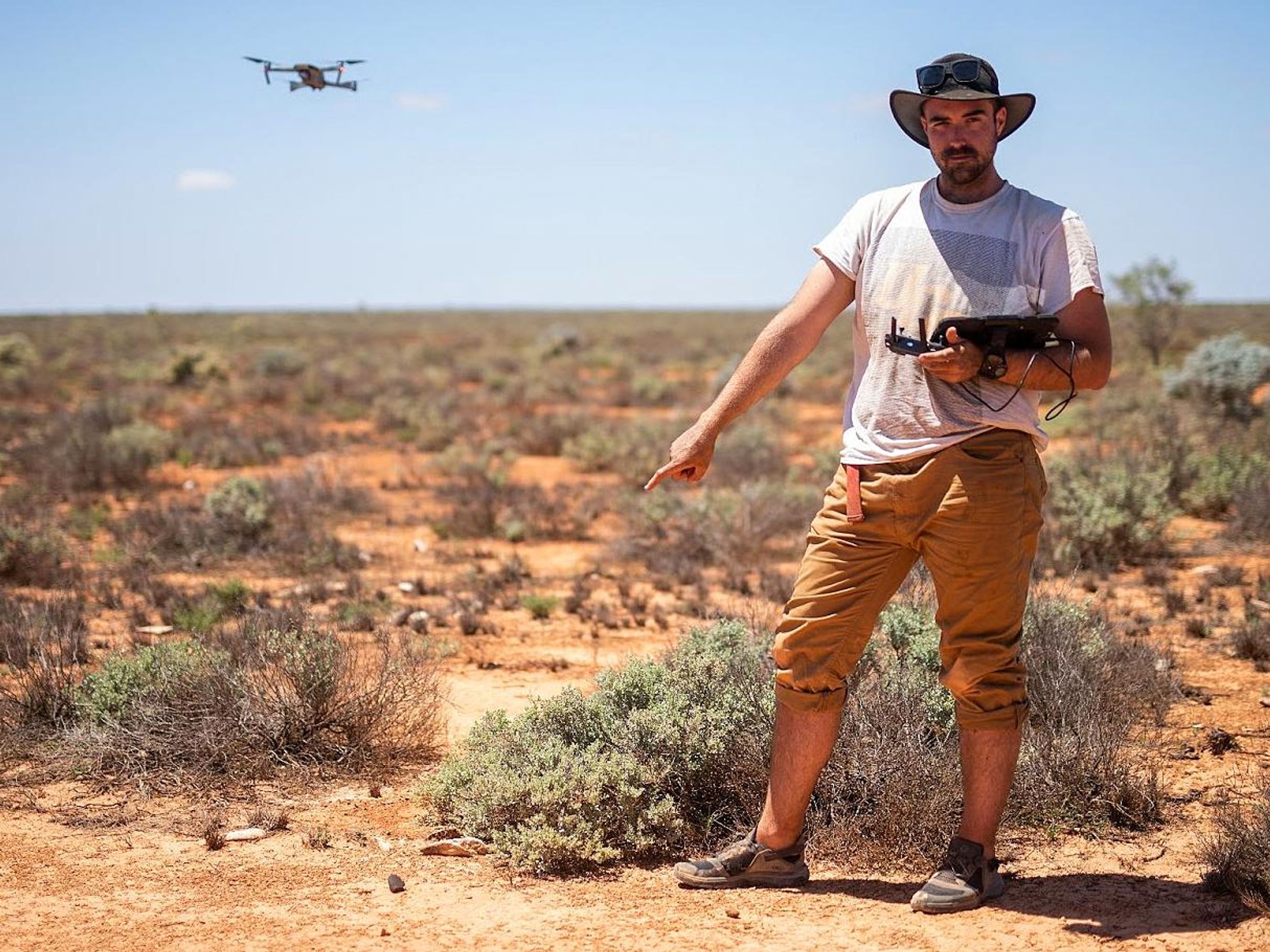 A man stands in the desert holding a remote controller for a drone that hovers in the air behind him. He is pointing to a small dark object on the ground.