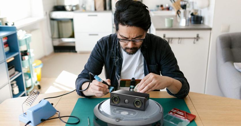 A man uses a soldering iron to work on an iRobot Create 3 robot, which has a Roomba-like body with a camera module mounted on top.