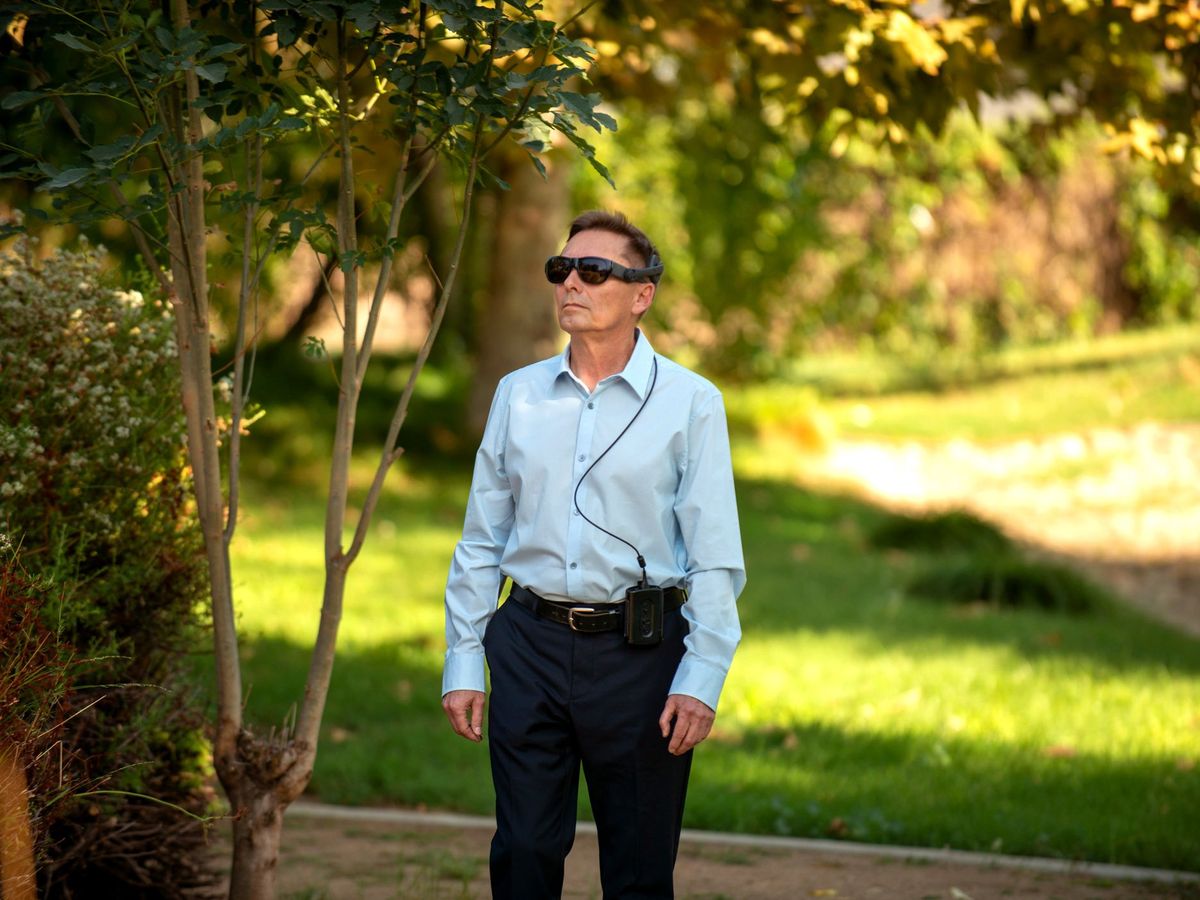 a man wearing a button-down shirt and pants walking through a park with tinted sunglasses and a wire coming down his shirt to a device hanging on his pants