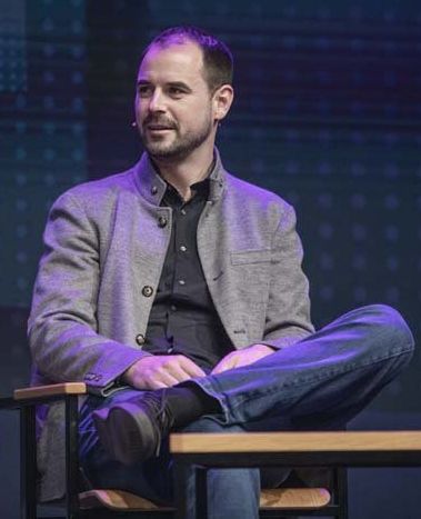 a man wearing a gray jacket and jeans sits in a chair.