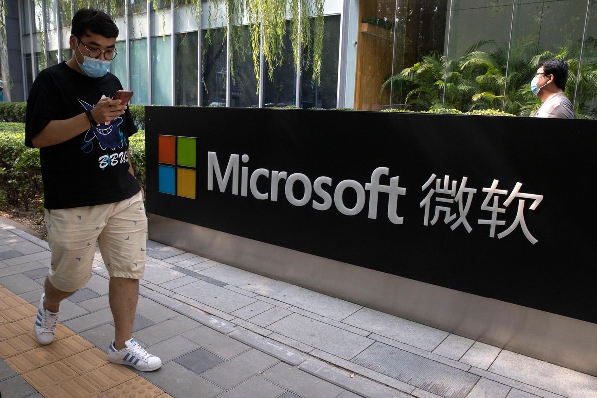 A man wearing a surgical mask holding a smartphone walks by a sign that says Microsoft and presumably “Microsoft” in Chinese characters as well