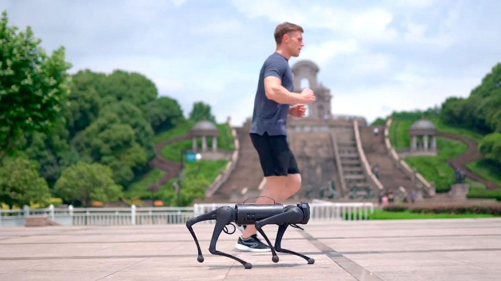 A man wearing athletic outfit jogs next to a Unitree Go1 quadruped robot on a park.