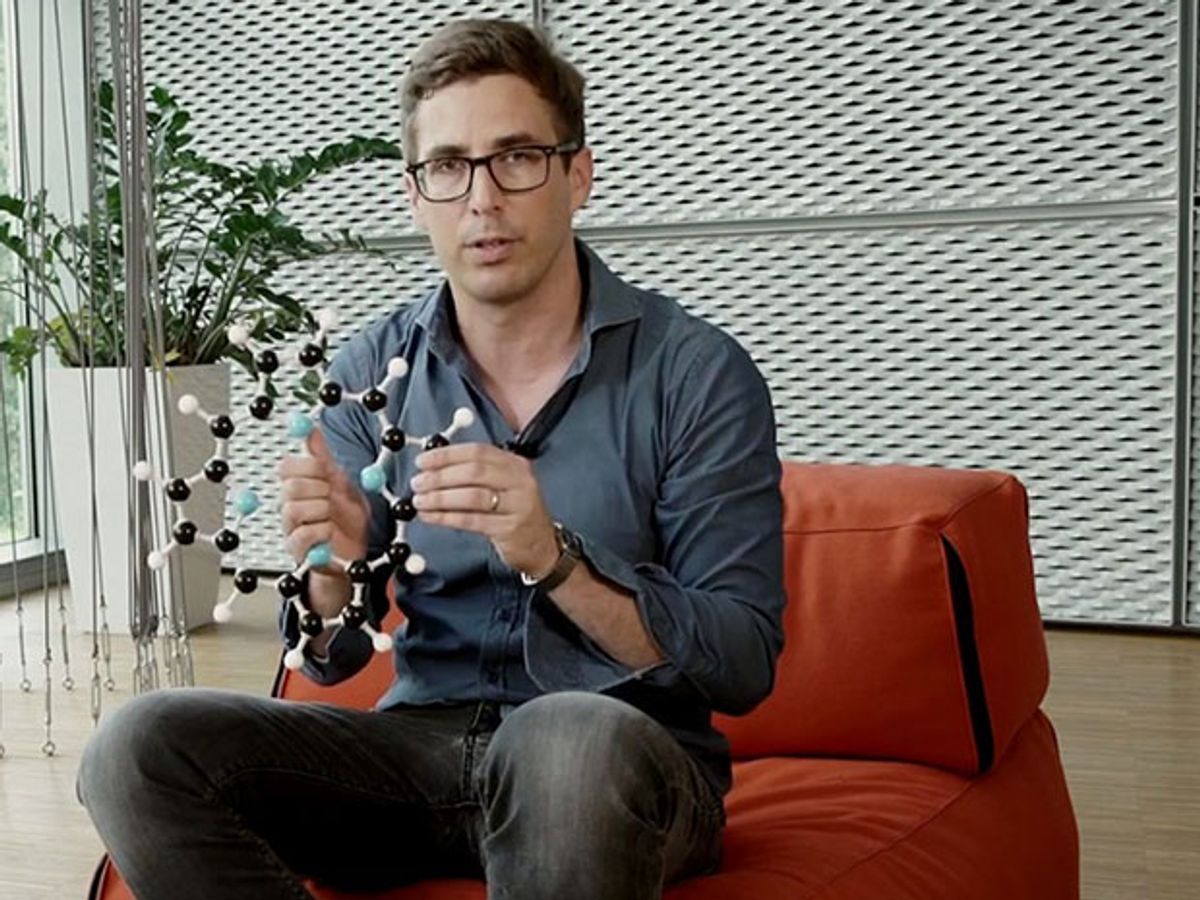 A man wearing black jeans and a grey-blue shirt sits holding a model of a ring-shaped molecule